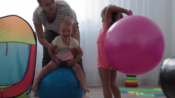 Barndom, Moderskap, spel, uppfostran och utveckling koncept - allmän plan för små barn pojke och flicka i samma ålder med ung mamma är glatt hoppa på rosa fitball och le i lekrum — Stockvideo