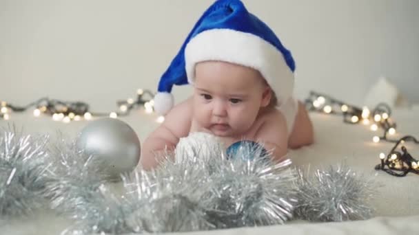 Merry christmas christmas and happy new year, infants, childhood, holidays concept - close-up naked 6 month old newborn baby in santa claus hat on his tummy crawls with decorations on christmas tree. — Stock Video
