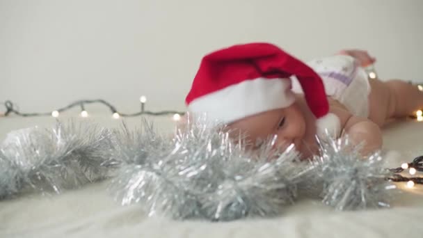 Vrolijk kerstfeest en gelukkig nieuwjaar, baby 's, jeugd, vakantie concept - close-up naakt 6 maanden oud pasgeboren baby in kerstman hoed op zijn buik kruipt met decoraties op kerstboom. — Stockvideo