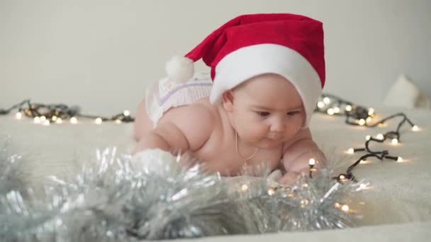 Boldog karácsonyt és boldog új évet, csecsemők, gyermekkor, ünnepek koncepció - közelkép meztelen 6 hónapos újszülött baba télapó claus kalap hasán kúszik díszek karácsonyfa. — Stock videók