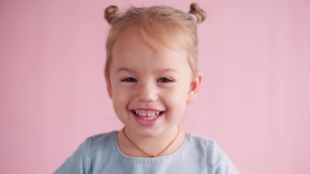 Enfance, célébration, anniversaire, concept de jeux - gros plan heureuse petite fille aux cheveux blonds en robe bleue regardant la caméra couvre les oreilles cris et montrant différentes émotions sur fond solide rose. — Video