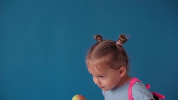 Childhood, school, education, upbringing, science concept - close-up happy little blonde caucasian slavic girl with backpack holding book and eats green apple smiling broadly on solid blue background — Stock Video