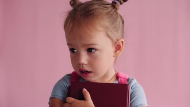 Kindertijd, school, onderwijs, opvoeding, wetenschap concept - close-up gelukkig klein blond Kaukasisch slavisch meisje in blauwe jurk met rugzak knuffels boek in handen brede glimlach op solide roze achtergrond — Stockvideo