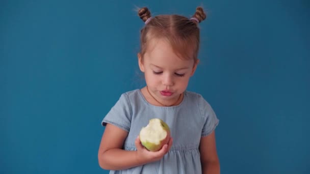 Dzieciństwo, szkoła, edukacja, wychowanie, koncepcja naukowa - zbliżenie szczęśliwa mała blondynka kaukaska słowiańska dziewczyna trzyma książkę i je zielone jabłko uśmiechnięte szeroko na solidnym niebieskim tle chroma klucz — Wideo stockowe