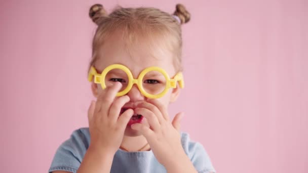 Childhood, celebration, birthday, games concept - close-up little blond girl in blue dress looking at camera and showing different emotions waving finger in yellow toy glasses on pink solid background — Stock Video