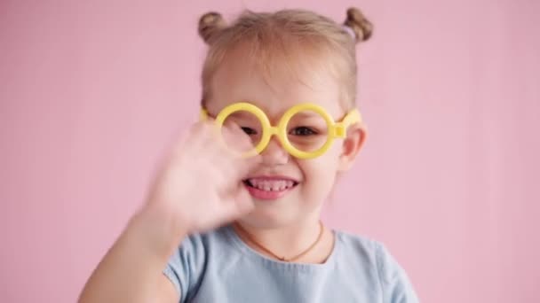 Dzieciństwo, uroczystość, urodziny, koncepcja gier - zbliżenie mała blondynka w niebieskiej sukience patrząc przed kamerą i pokazując różne emocje machając ręką w żółtych okularach zabawki na różowym solidnym tle. — Wideo stockowe