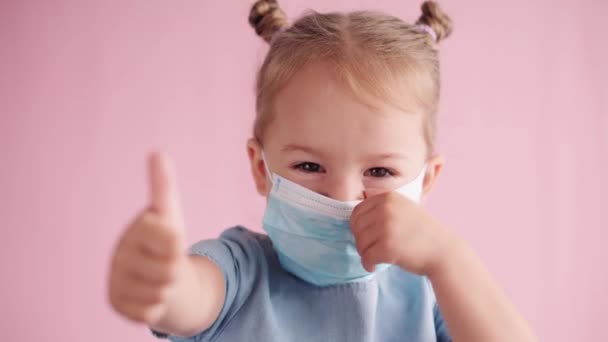 Quarantine, covid-19, safe distance, distance and online learning, school concept - close-up of little blonde girl in blue medical mask demonstrates different emotions on solid pink background. — Stock Video