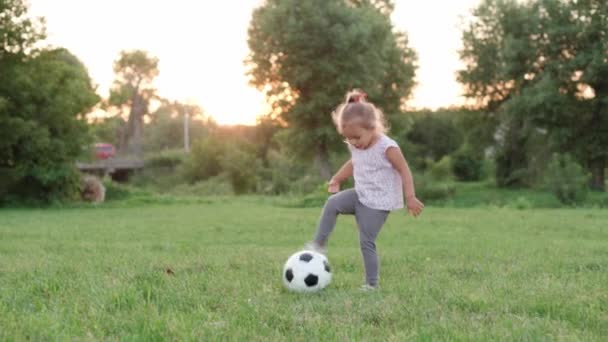 Infancia, juegos y entretenimiento, deportes, cultura física, parques y concepto al aire libre - niña pequeña rubia jugar al fútbol con bola blanca y negra en el césped verde brillante en el parque de verano al atardecer. — Vídeo de stock