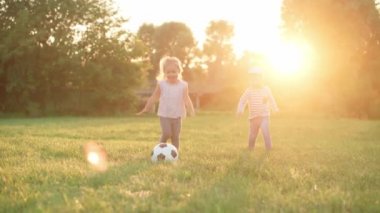 Çocukluk, oyunlar ve eğlence, spor, fiziksel kültür, parklar ve açık hava konsepti - küçük çocuklar ve kızlar gün batımında parkta açık yeşil çimlerde siyah ve beyaz topla futbol oynuyorlar..