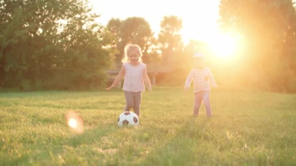 Infância, jogos e entretenimento, esportes, cultura física, parques e conceito ao ar livre - meninos e meninas crianças jogam futebol com bola preta e branca no gramado verde brilhante no parque ao pôr do sol. — Vídeo de Stock