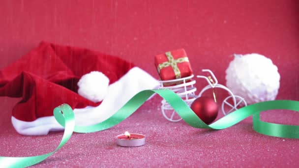クリスマス、冬、幸せな新年のコンセプト-白い雪で覆われた赤いクリスマスの背景は、トランクの贈り物と燃えるキャンドルやおもちゃの自転車でサンタの帽子に落ちます。フラット、トップビュー、オーバーヘッドループビデオ — ストック動画