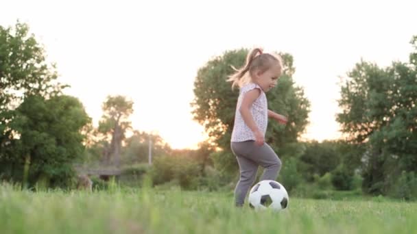 Infância, jogos e entretenimento, esportes, cultura física, conceito ao ar livre - menina loira criança andando em círculo em torno de bola preta e branca e cair no gramado verde no pôr do sol do parque de verão. — Vídeo de Stock