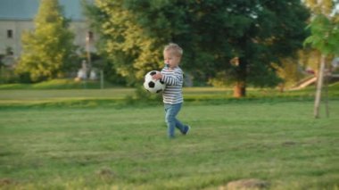 Çocukluk, oyunlar ve eğlence, spor, fiziksel kültür, açık hava konsepti - küçük sarışın çocuk 2 yıl koştu ve elinde tuttuğu siyah beyaz top yaz parkında güneş batarken açık yeşil çimlerde.