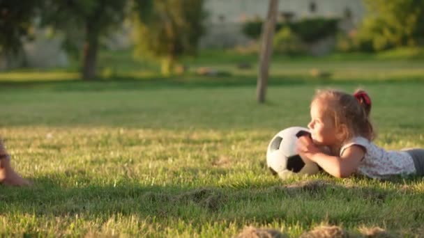 Infância, jogos e entretenimento, esportes, cultura física, parques e conceito ao ar livre - menina criança feliz com a jovem mãe jogar futebol com bola preta e branca no gramado verde no parque ao pôr do sol. — Vídeo de Stock