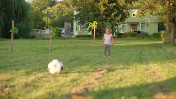 Dzieciństwo, gry i rozrywka, sport, kultura fizyczna, parki i koncepcja plenerowa - mała blondynka gra w piłkę nożną z czarno-białą piłką na jasnozielonym trawniku w letnim parku o zachodzie słońca. — Wideo stockowe