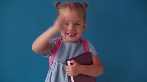 Childhood, school, education, upbringing, science concept - close-up happy little blonde caucasian slavic girl with backpack holding book and waving goodbye smiling broadly on solid blue background — Stock Video