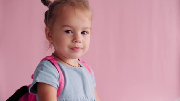 Childhood, school, education, upbringing, science concept - close-up happy little blonde caucasian slavic girl in blue dress with backpack flirts closes hands makes air kiss on solid pink background — Stock Video