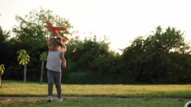Infância, jogos e entretenimento, esportes, cultura física, parques e conceito ao ar livre - criança pequena menina loira segurando e lançando avião de brinquedo laranja no gramado verde brilhante no parque de verão ao pôr do sol. — Vídeo de Stock