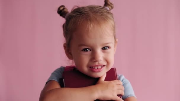 Infância, escola, educação, educação, conceito de ciência - close-up feliz pequena menina eslava caucasiana loira em vestido azul com abraços mochila livro em mãos sorrindo amplamente no fundo rosa sólido — Vídeo de Stock