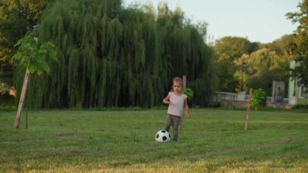 Infância, jogos e entretenimento, esportes, cultura física, parques e conceito ao ar livre - menina loira criança jogar futebol com bola preta e branca no gramado verde brilhante no parque de verão ao pôr do sol. — Vídeo de Stock