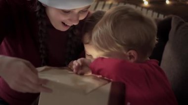 Xmas, kış, yeni yıl, kutlama, aile konsepti - iki küçük çocuklu mutlu anne ve kırmızı elbiseli kız hediye kutusu açık, süslü Noel ağacının yanında oturuyor. Parlak bir ışık.
