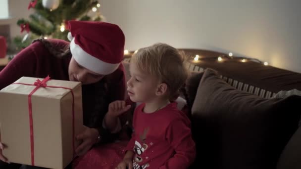 Vánoce, zima, Nový rok, Oslava, rodinný koncept - Mladá šťastná matka se dvěma malými dětmi chlapec a dívka oblečeni ve vánočním oblečení otevřené dárkové krabice sedí na pohovce u zdobeného stromu večer — Stock video
