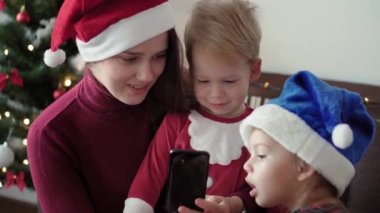 Xmas, kış, yeni yıl, kutlama, aile konsepti Noel kıyafetleri ve Noel Baba şapkası giymiş mutlu bir aile online alışveriş yapıyor akıllı telefon ve kredi kartı takılı ağacın yanında oturuyor..