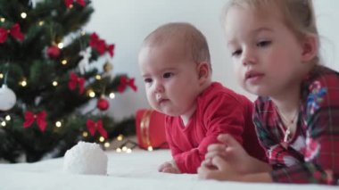 Mutlu Noeller ve mutlu yıllar, bebek, çocukluk, tatil konsepti - yakın plan 6 aylık Noel Baba şapkalı bebek ve ablası süsleme balosu yılbaşı ağacı ile sürünen kırmızı tulum