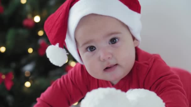 Feliz Navidad y feliz año nuevo, bebés, infancia, concepto de vacaciones - primer plano bebé recién nacido de 6 meses en sombrero de Santa Claus y traje de cuerpo rojo en su barriga se arrastra con bolas de decoración en el árbol de Navidad — Vídeo de stock