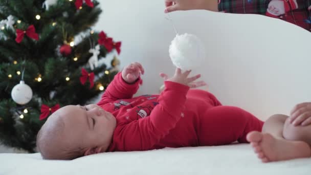Καλά Χριστούγεννα και ευτυχισμένο το νέο έτος, βρέφος, παιδική ηλικία, έννοια των διακοπών - close-up 6 μηνών νεογέννητο μωρό σε Σάντα Claus καπέλο και κόκκινο bodysuit με μεγαλύτερη αδελφή σέρνεται με διακοσμήσεις μπάλα χριστουγεννιάτικο δέντρο — Αρχείο Βίντεο