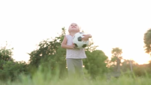 Childhood, games and entertainment, sports, physical culture, open air concept - happy little kid blonde girl carries in hands black and white ball and points finger up at sky on in summer park sunset — Stock Video