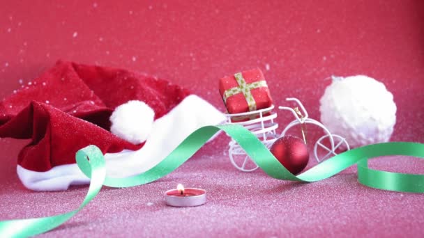 Navidad, invierno, concepto de año nuevo - fondo rojo de Navidad cubierto de nieve blanca Santa sombrero con vela ardiente y bicicleta de juguete con regalo figurado en el tronco. reorientar el plan de lejos hacia el primer plano — Vídeos de Stock