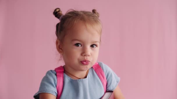 Childhood, school, education, upbringing, science concept - close-up little blonde caucasian slavic girl in blue dress with backpack holds book and sternly waves index finger on solid pink background — Stock Video