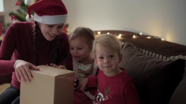 Kerstmis, winter, Nieuwjaar, Feestdag, Familie concept - Jonge gelukkige moeder met twee kleine kinderen jongen en meisje gekleed in kerstkleding open geschenkdoos zittend op bank in de buurt van versierde boom 's avonds — Stockvideo