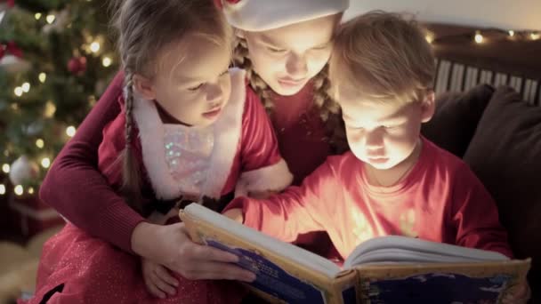 Xmas, kış, yeni yıl, kutlama, aile konsepti - Genç mutlu anne, iki küçük çocuk, kırmızı elbiseli kız ve erkek çocuk, peri masalları okuyun, süslenmiş yılbaşı ağacının yanında, kitaptan parlak bir ışık — Stok video