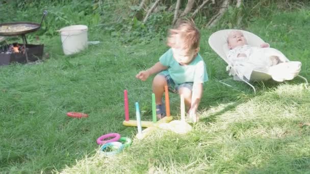 Infância, infância, piquenique, jogos educativos, desenvolvimento, conceito de férias de verão - menino jogar plástico Ring Toss Quoits no jardim. bebê recém-nascido deitado em cadeira de balanço fundo de grama verde — Vídeo de Stock