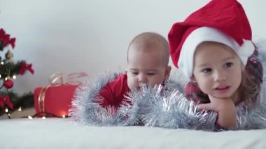 Mutlu Noeller ve mutlu yıllar, bebek, çocukluk, tatil konsepti - yakın plan 6 aylık Noel Baba şapkalı bebek ve ablası süsleme balosu yılbaşı ağacı ile sürünen kırmızı tulum