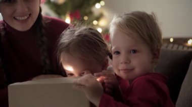 Xmas, kış, yeni yıl, kutlama, aile konsepti - iki küçük çocuklu mutlu anne ve kırmızı elbiseli kız hediye kutusu açık, süslü Noel ağacının yanında oturuyor. Parlak bir ışık.