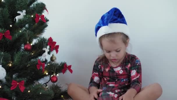 Xmas, vinter, nytt år, fest, familj koncept - glad smide liten flicka klädd i julkläder leker med dekorationer, kransar och visar känslor sitter nära dekorerade träd på kvällen — Stockvideo