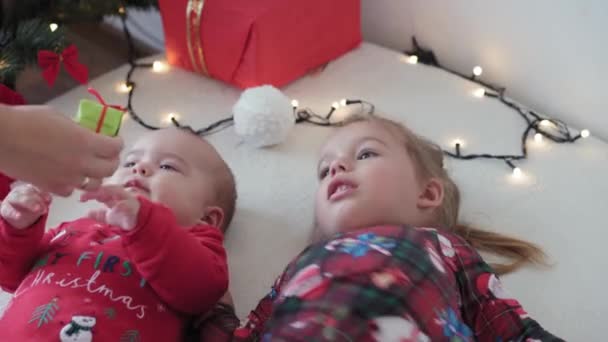 Весёлого Рождества и счастливого Нового года, младенец, детство, праздники концепция - крупным планом 6-месячный новорожденный ребенок в шляпе Санта-Клауса и красном боди-костюме со старшей сестрой лежал на кровати с шарами возле рождественской елки. — стоковое видео