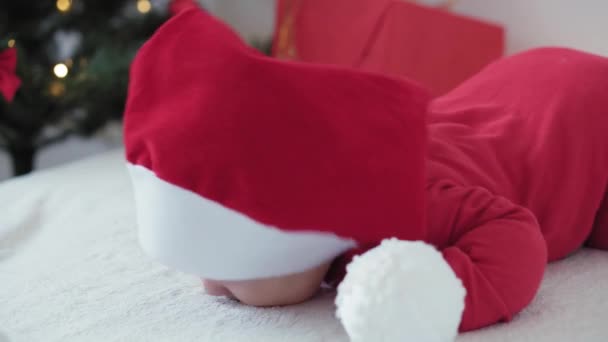 Merry xmas and happy new year, infants, childhood, holidays concept - close-up 6 month old newborn baby in santa claus hat and red bodysuit on his tummy crawls with decorations balls on christmas tree — Stock Video