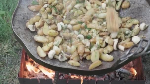 Cafés y restaurantes, picnic, concepto de cocina oriental primer plano agitar patatas y setas espátula de madera en una sartén enorme para cocinar alimentos sobre fogatas al aire libre: carne, verduras otros platos. — Vídeo de stock