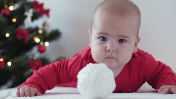 Καλά Χριστούγεννα και ευτυχισμένο το νέο έτος, τα βρέφη, την παιδική ηλικία, τις διακοπές έννοια - close-up 6 μηνών νεογέννητο μωρό σε Σάντα Claus καπέλο και κόκκινο bodysuit στην κοιλιά του σέρνεται με μπάλες διακόσμησης στο χριστουγεννιάτικο δέντρο — Αρχείο Βίντεο