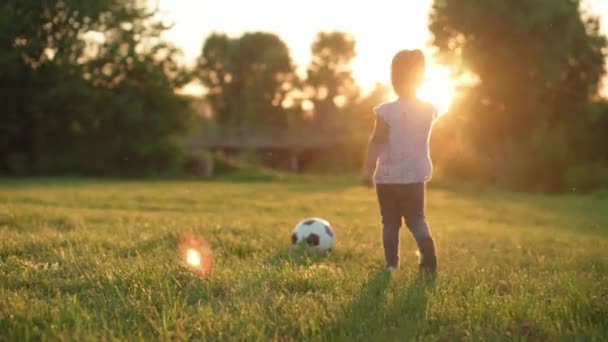 Infância, jogos e entretenimento, esportes, cultura física, parques e conceito ao ar livre - menina loira criança jogar futebol com bola preta e branca no gramado verde brilhante no parque de verão ao pôr do sol. — Vídeo de Stock