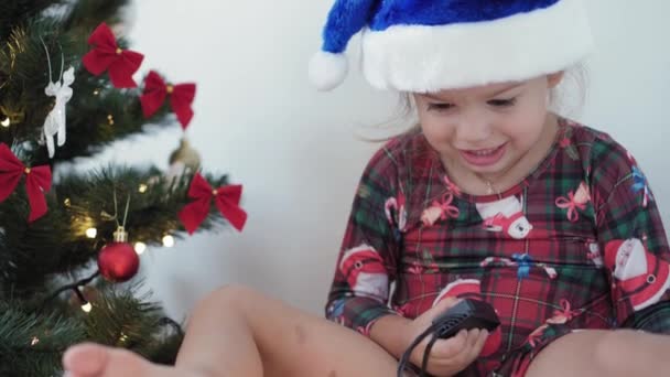 Kerstmis, winter, Nieuwjaar, Feest, Familie concept - gelukkig lachend meisje gekleed in kerstkleding speelt met decoraties, bloemenslingers en toont emotie zitten in de buurt van versierde boom in de avond — Stockvideo