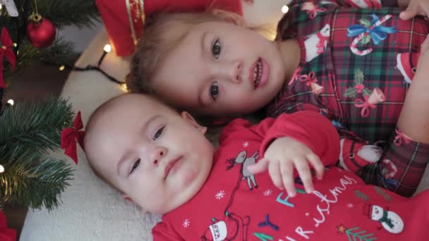 Merry xmas and happy new year, infant, childhood, holidays concept - close-up 6 month old newborn baby in santa claus hat and red bodysuit with elder sister lay on bed with balls near christmas tree. — Stock Video