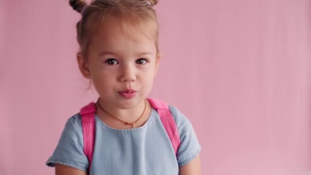 Childhood, school, education, upbringing, science concept - close-up little blonde caucasian slavic girl with backpack holding book and showing discontent and disagreement on solid pink background — Stock Video