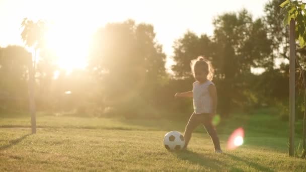 Infância, jogos e entretenimento, esportes, cultura física, parques e conceito ao ar livre - menina loira criança jogar futebol com bola preta e branca no gramado verde brilhante no parque de verão ao pôr do sol. — Vídeo de Stock