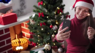 Xmas, kış, yeni yıl, kutlama, aile konsepti - Noel Baba şapkalı kız uzaktan iyi bayramlar diler video konferansı akıllı telefon online dalga maytapları ile karantinada Noel ağacının yanında oturuyor