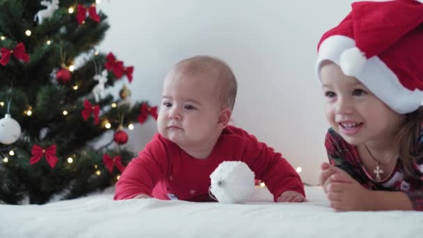 Merry xmas and happy new year, infant, childhood, holidays concept - close-up 6 month old newborn baby in santa claus hat and red bodysuit with elder sister crawls with decorations ball christmas tree — Stock Video